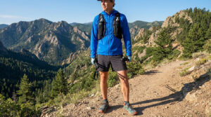 Un homme en tenue de trail bleue et short noir, avec sac d'hydratation et bâtons, pose sur un sentier de montagne ensoleillé.